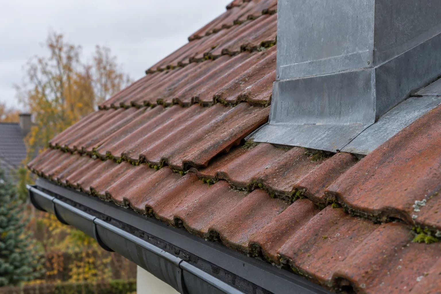 Dach z ceramicznych dachówek przy kominie z blachy. Jedna dachówka jest lekko przesunięta.